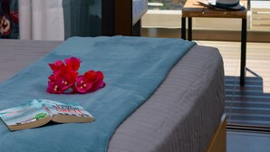 A bedroom with a bed, a window with a view of the sea, and a small table with a hat and book. - Villa Olga (Agios Nikolaos)