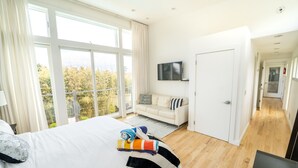 A modern hotel room with a large bed, a flat-screen TV, a sofa, and a view of trees through large windows.