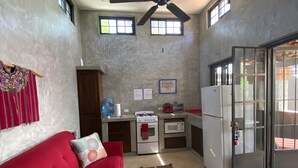 Un espacio habitable compacto con un sofá rojo, una mesita, ventilador de techo, kitchenette y puerta corrediza de vidrio.