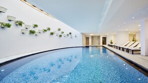 Una piscina interior moderna con superficie de agua azul cristalina, rodeada de sillones de descanso y plantas en macetas en las paredes.