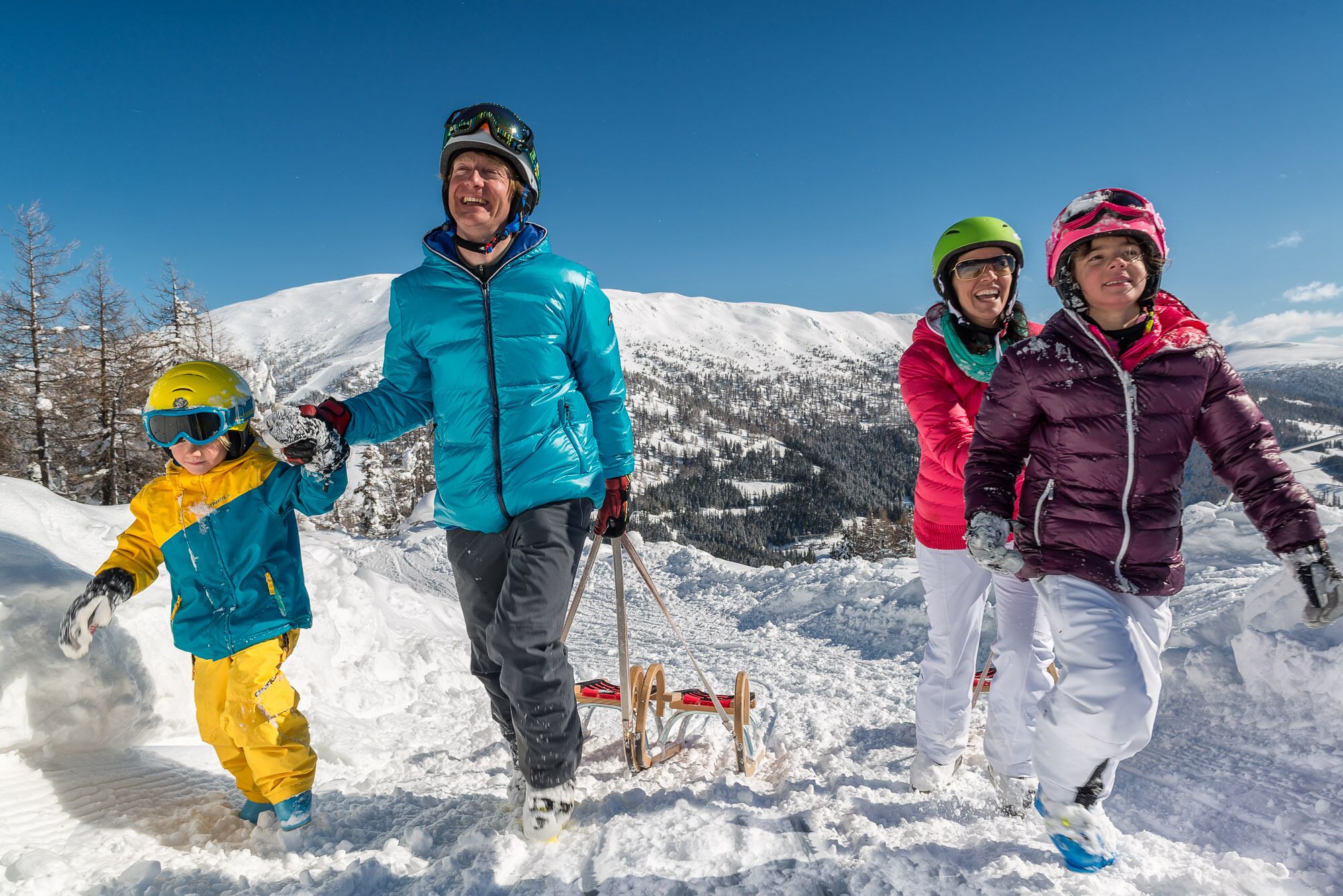 A family of four on a snowy mountain slope, dressed in winter sports gear, with a clear sky and snow-covered landscape in the background.