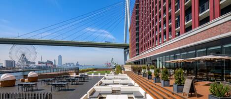 A rooftop terrace with outdoor seating, a view of a city skyline, and a modern building with red accents.