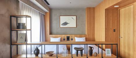 A modern hotel room with a wooden headboard, a large window with curtains, a wooden sideboard with decorative items, and a grey sofa with patterned cushions.