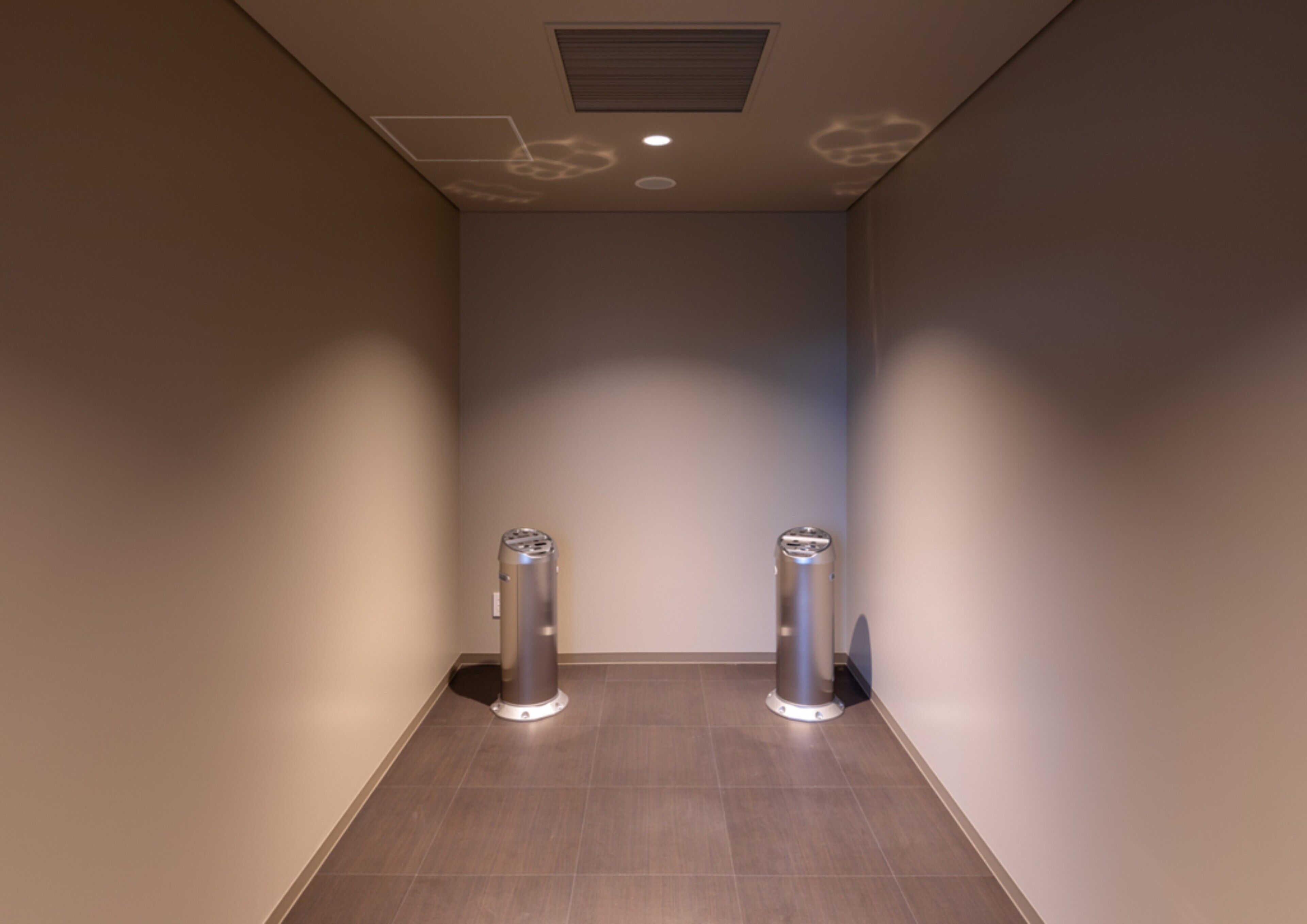 A narrow hallway with two cylindrical objects on the floor, a ceiling vent, and a recessed light fixture.