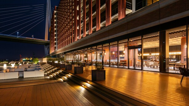 Een modern hotel met een overdekt terras, uitzicht op een brug, en een restaurant met terras.