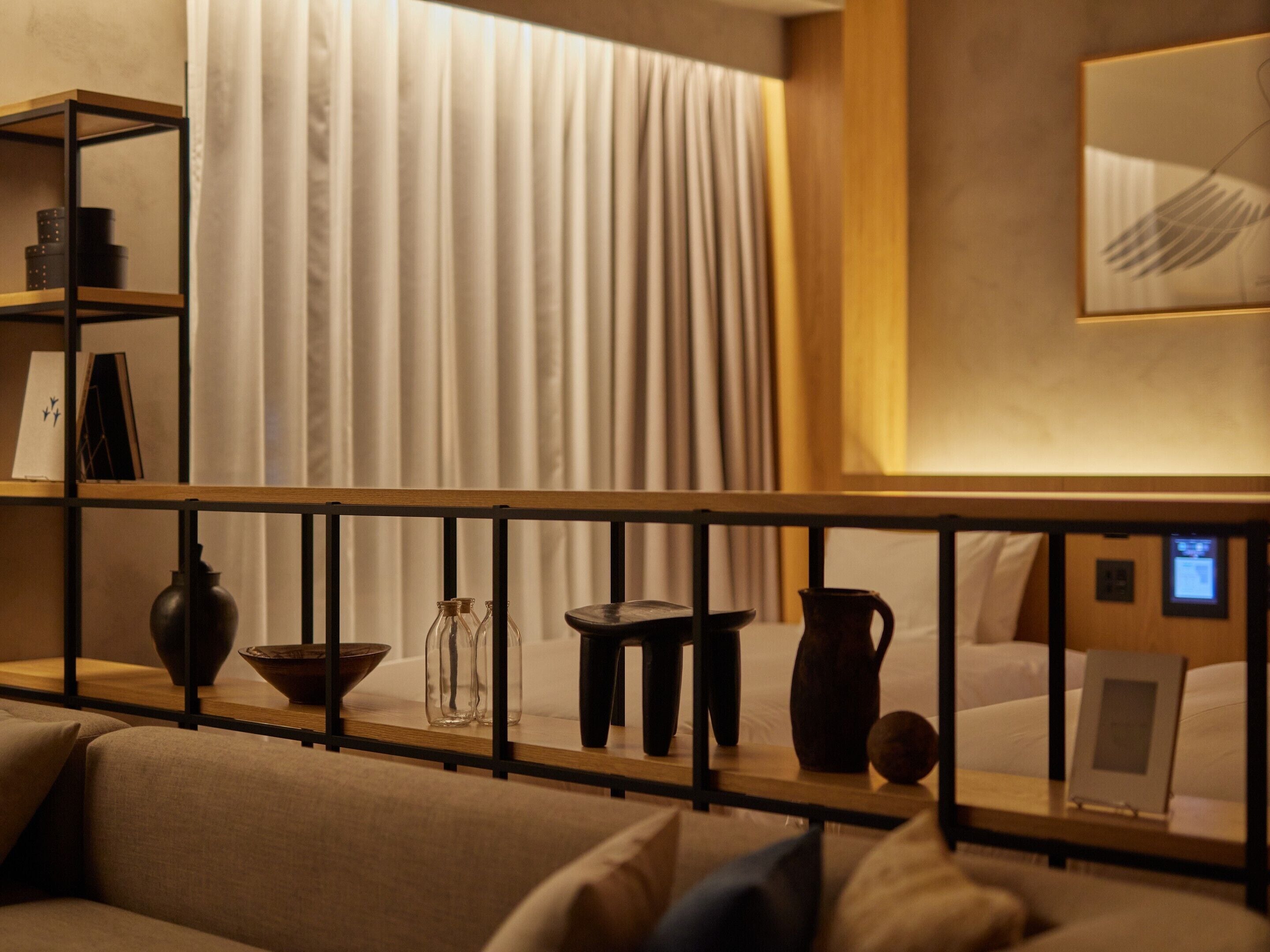 A modern hotel room with a sofa, a wooden shelf with decorative items, a bed with a digital control panel, and a large window with curtains.