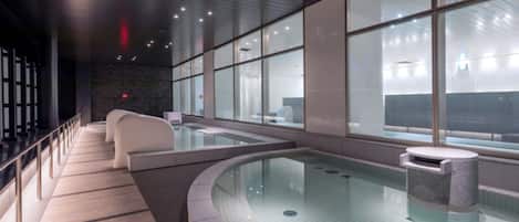 Un área de piscina interior moderna con paredes de vidrio transparente, una piscina de forma curva y un banco de piedra.