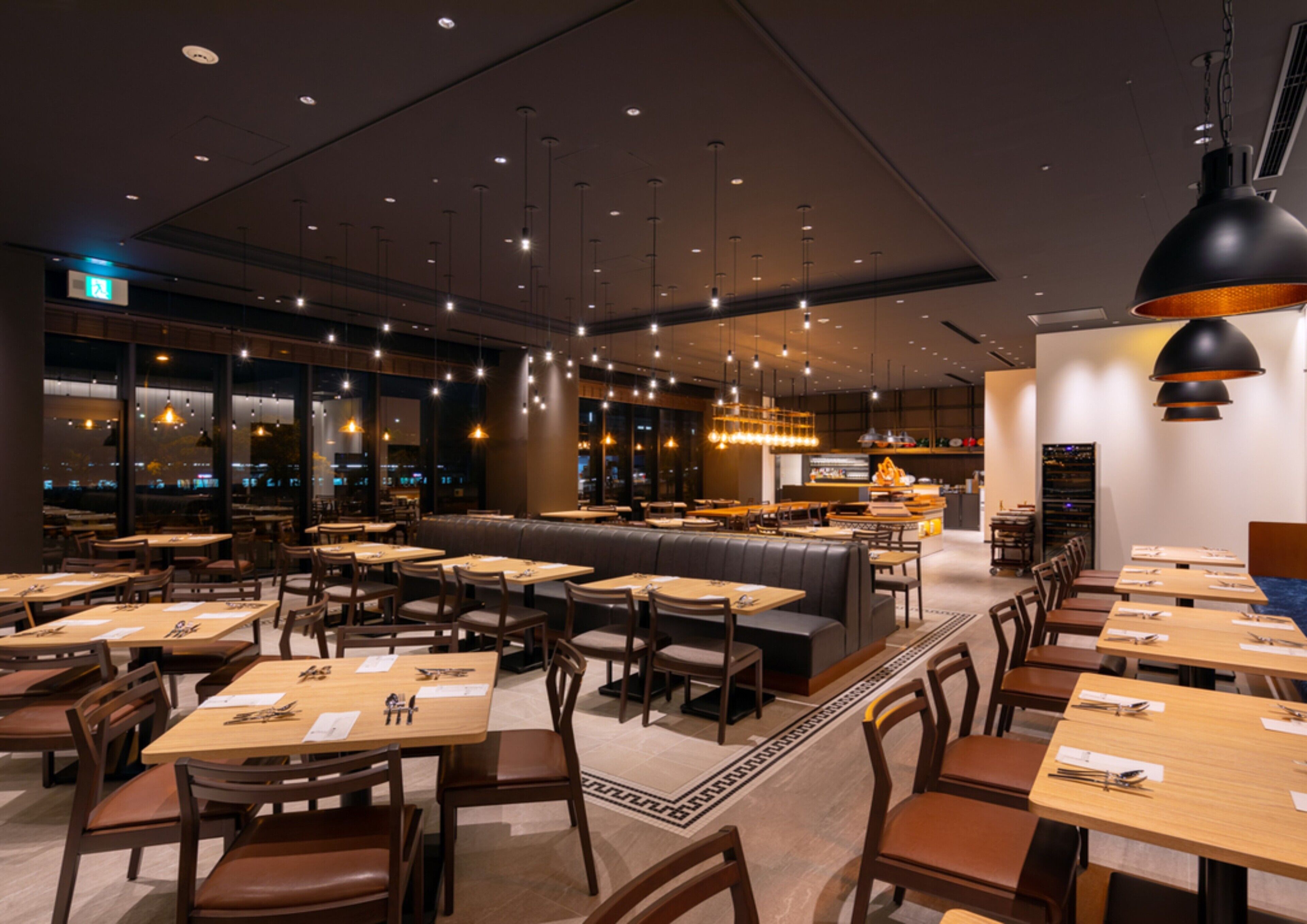 A modern restaurant interior with multiple dining tables and booths, overhead lighting, and a view of the city through large windows.