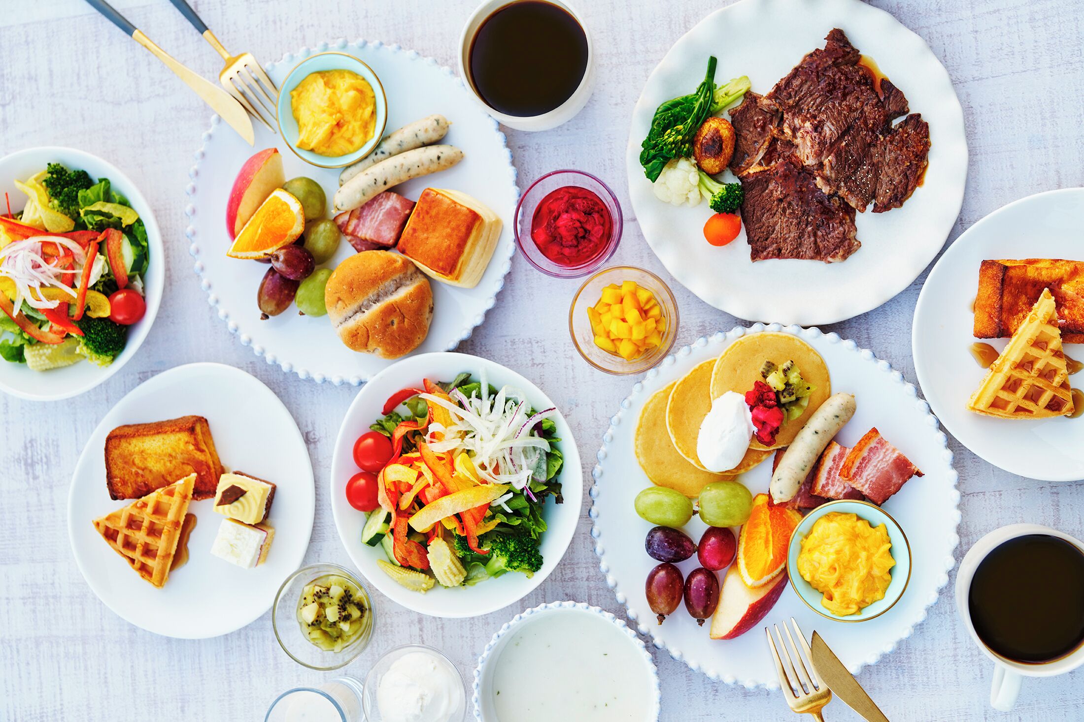 A table with various breakfast dishes including waffles, eggs, sausages, and salads.