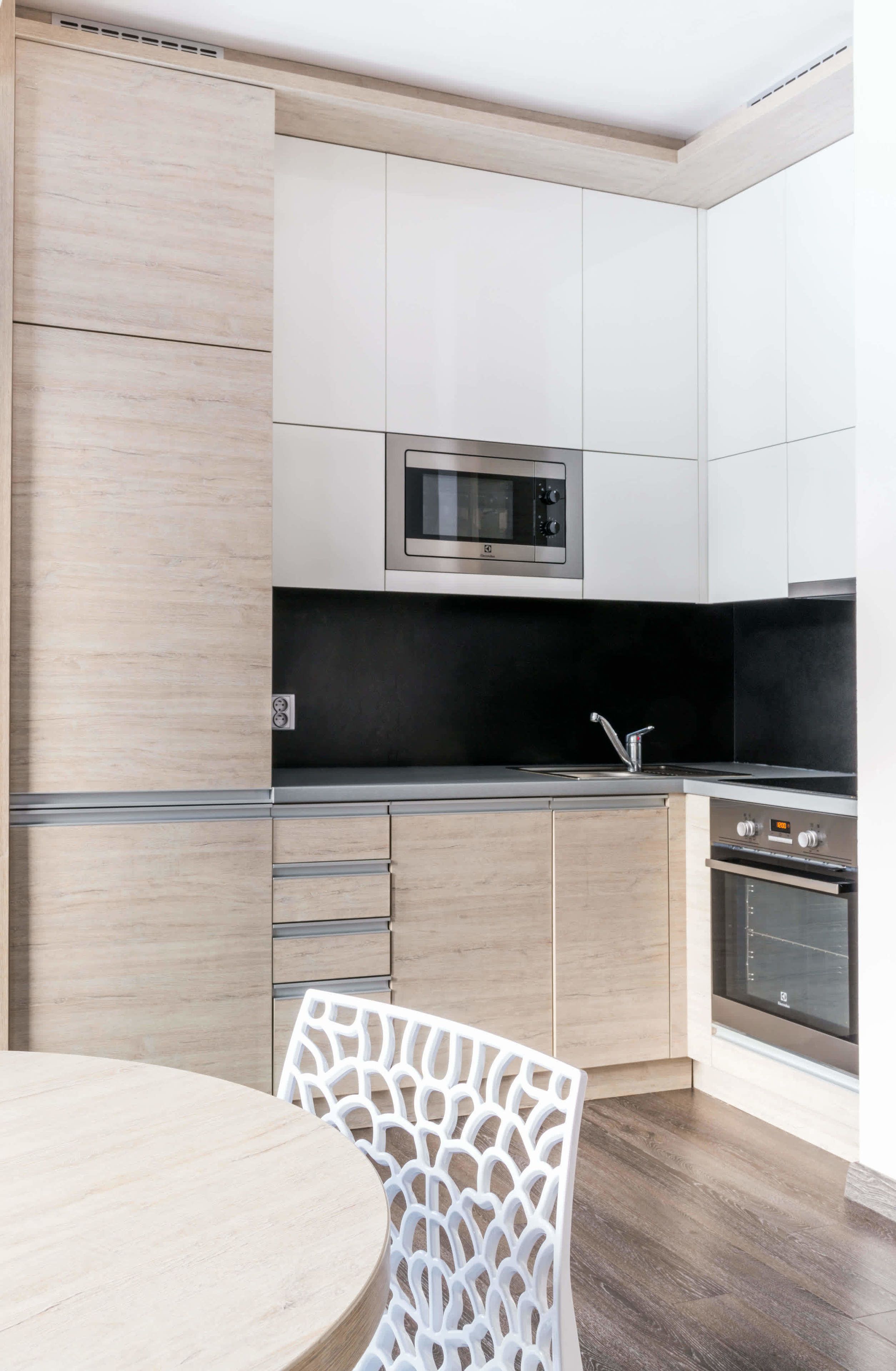 A modern kitchen with wooden cabinets, a stainless steel microwave, a black countertop, and a white chair.