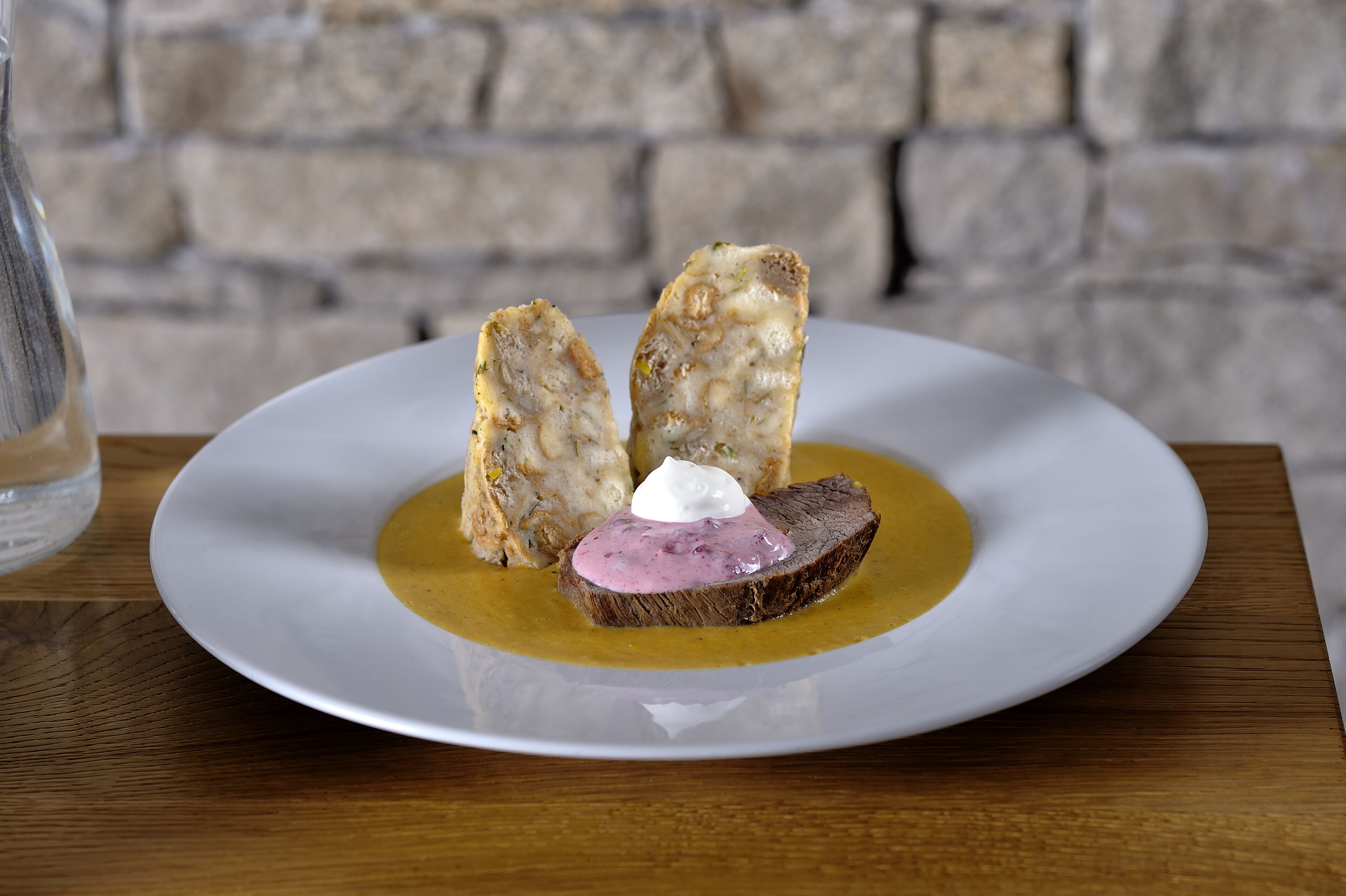 A white plate with a slice of meat topped with a dollop of sauce and a piece of bread, set on a wooden table against a stone wall background.
