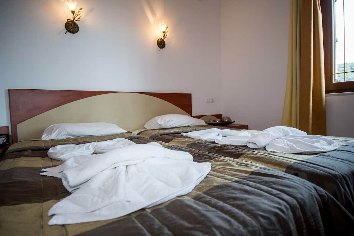 A hotel room with a bed, two wall sconces, a window with curtains, and a bedside table with a lamp.