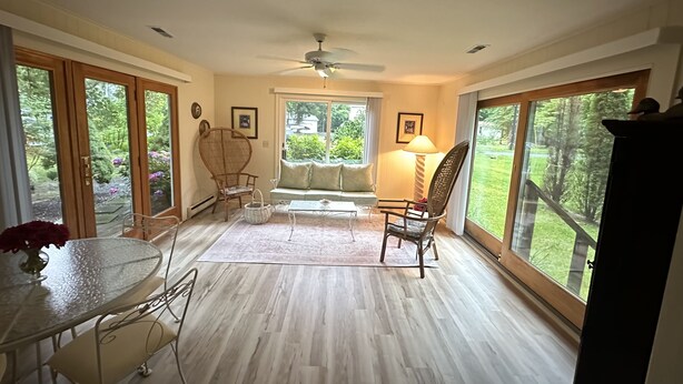 A spacious room with a ceiling fan, large windows, a dining table with chairs, a sofa, and a lamp. - Super Clean Home on a Quiet Cul-De-Sac in the Woods. (Lewes)