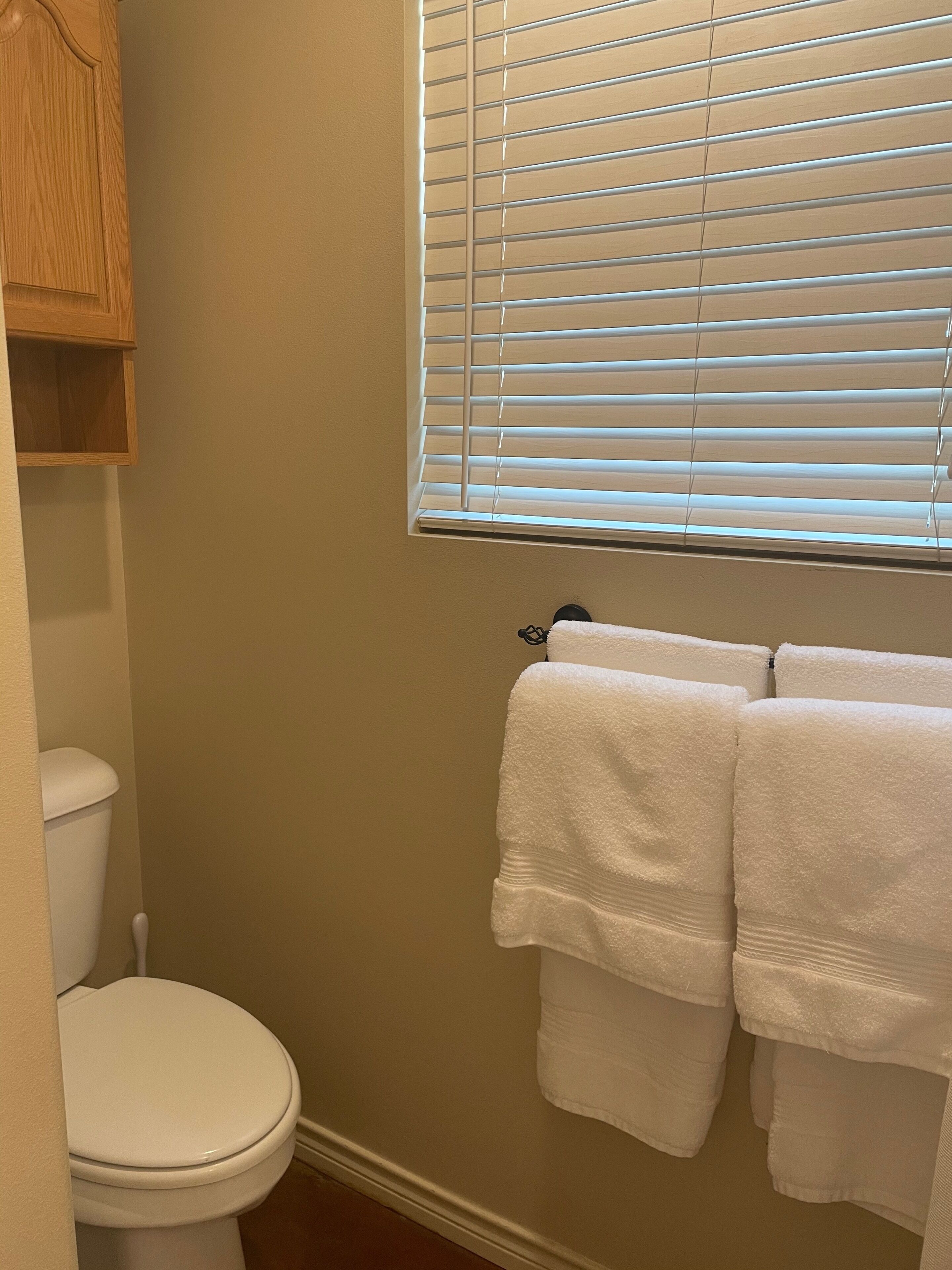 A bathroom with a toilet, a window with blinds, and two towels hanging on a rack.