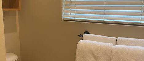 A bathroom with a toilet, a window with blinds, and two towels hanging on a rack.