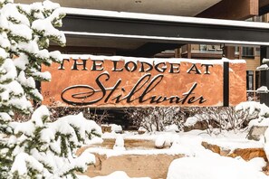 Ein Schild mit der Aufschrift "The Lodge at Stillwater" in rustikalem Design, umgeben von schneebedeckter Vegetation und Steinen.