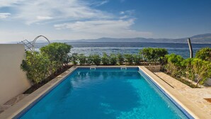 A rectangular swimming pool with clear blue water, surrounded by a tiled deck and low green shrubs. - Villa Violet (Supetar)