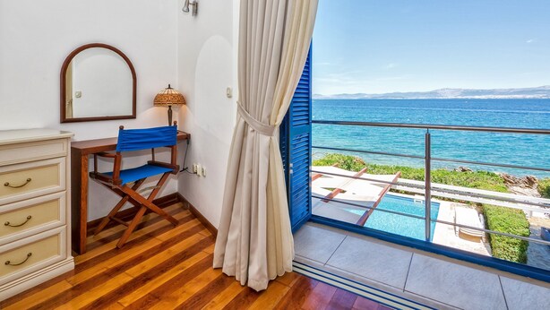A room with a wooden floor, a desk with a chair, a mirror, a lamp, and a large window with a view of the sea and a pool. - Villa Violet (Supetar)