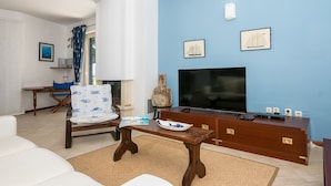 Ein Wohnzimmer mit einem weißen Sofa, einem Holztisch, einem blauen Sessel, einem Fernseher und einer Holzkommode.