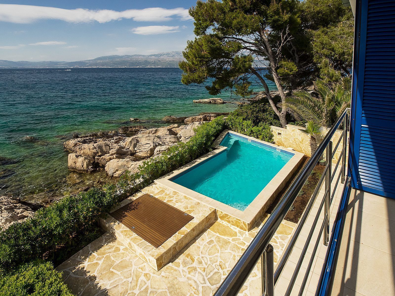Una piscina con agua azul cristalina, rodeada de baldosas de piedra y una terraza de madera, con vistas a un litoral rocoso y al mar.