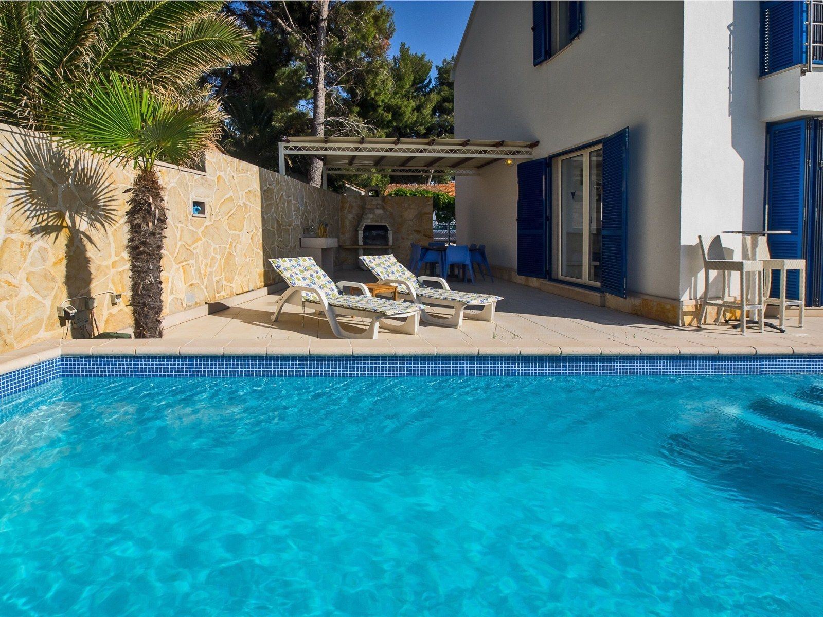 Ein Swimmingpool mit klarem, blauem Wasser, umgeben von einer Steinmauer und einem weißen Gebäude mit blauen Fensterläden. Liegestühle und eine Pergola mit Tisch und Stühlen sind ebenfalls vorhanden.