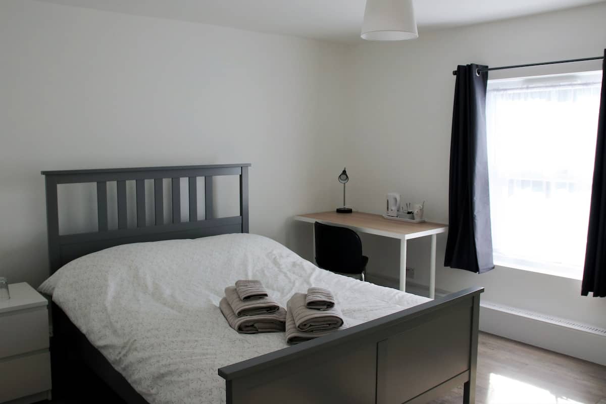 A neatly made bed with folded towels, a desk with a lamp, and a window with curtains.