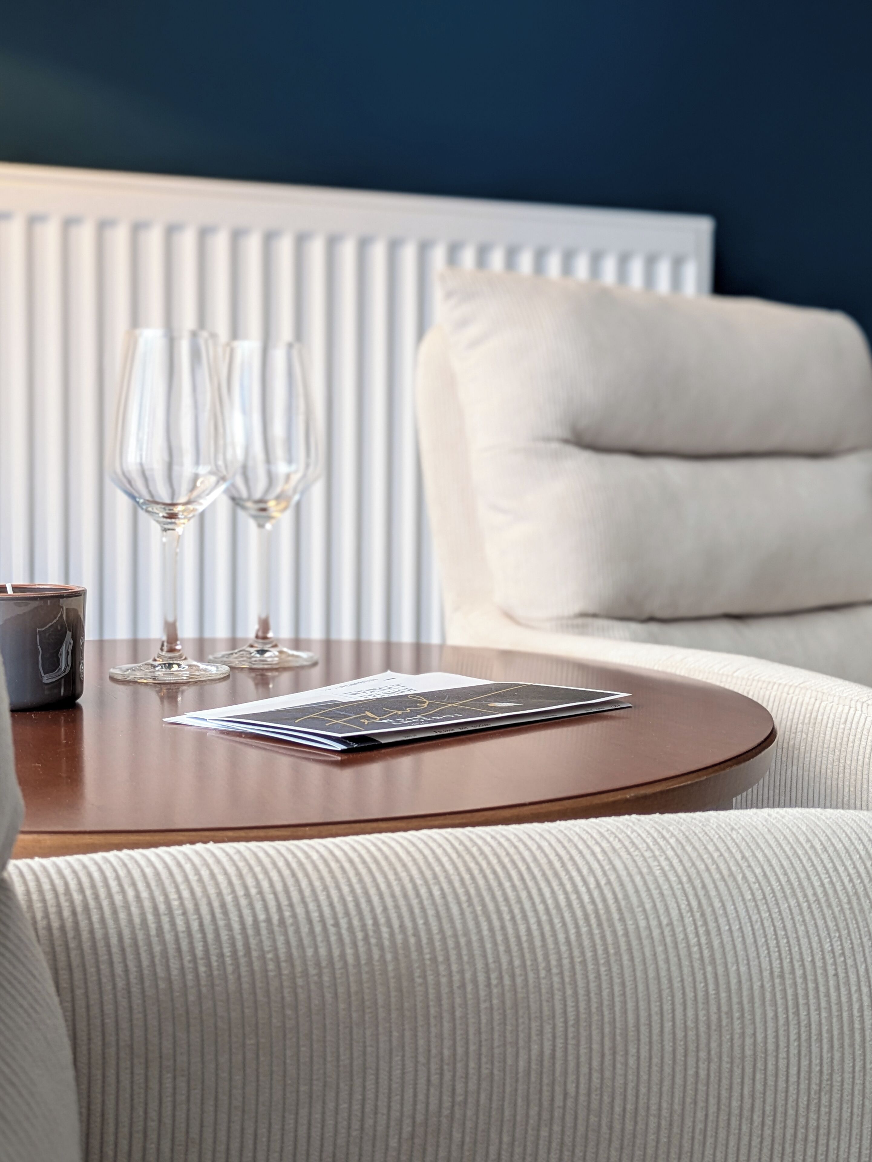 Ein runder Holztisch mit einer Zeitschrift, zwei Gläsern und einer Kerze. Im Hintergrund steht ein weißes, gepolstertes Sofa.