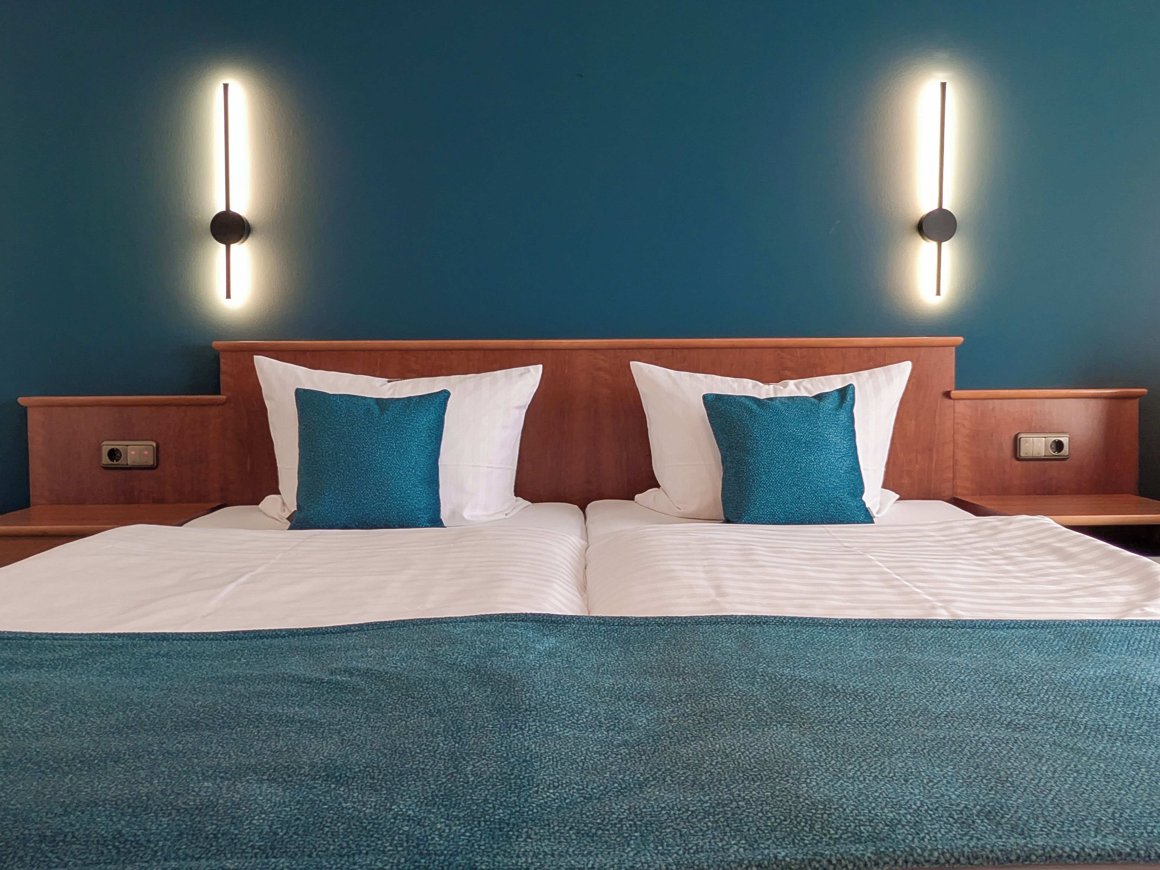 A double bed with white and teal bedding, wooden headboard, and two bedside tables with wall-mounted lights.