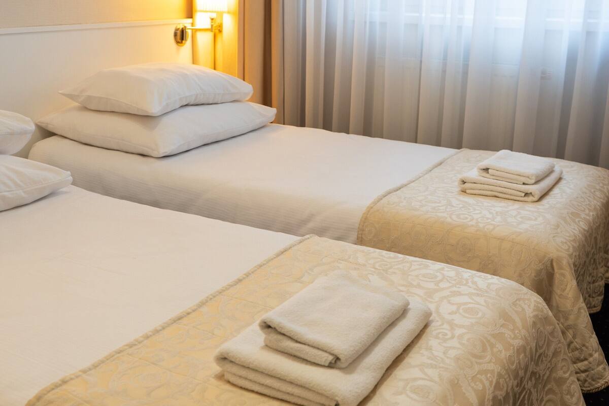 Habitación de hotel con dos camas, cada una con cubrecama y almohadas blancas, y un ventanal con cortinas traslúcidas.
