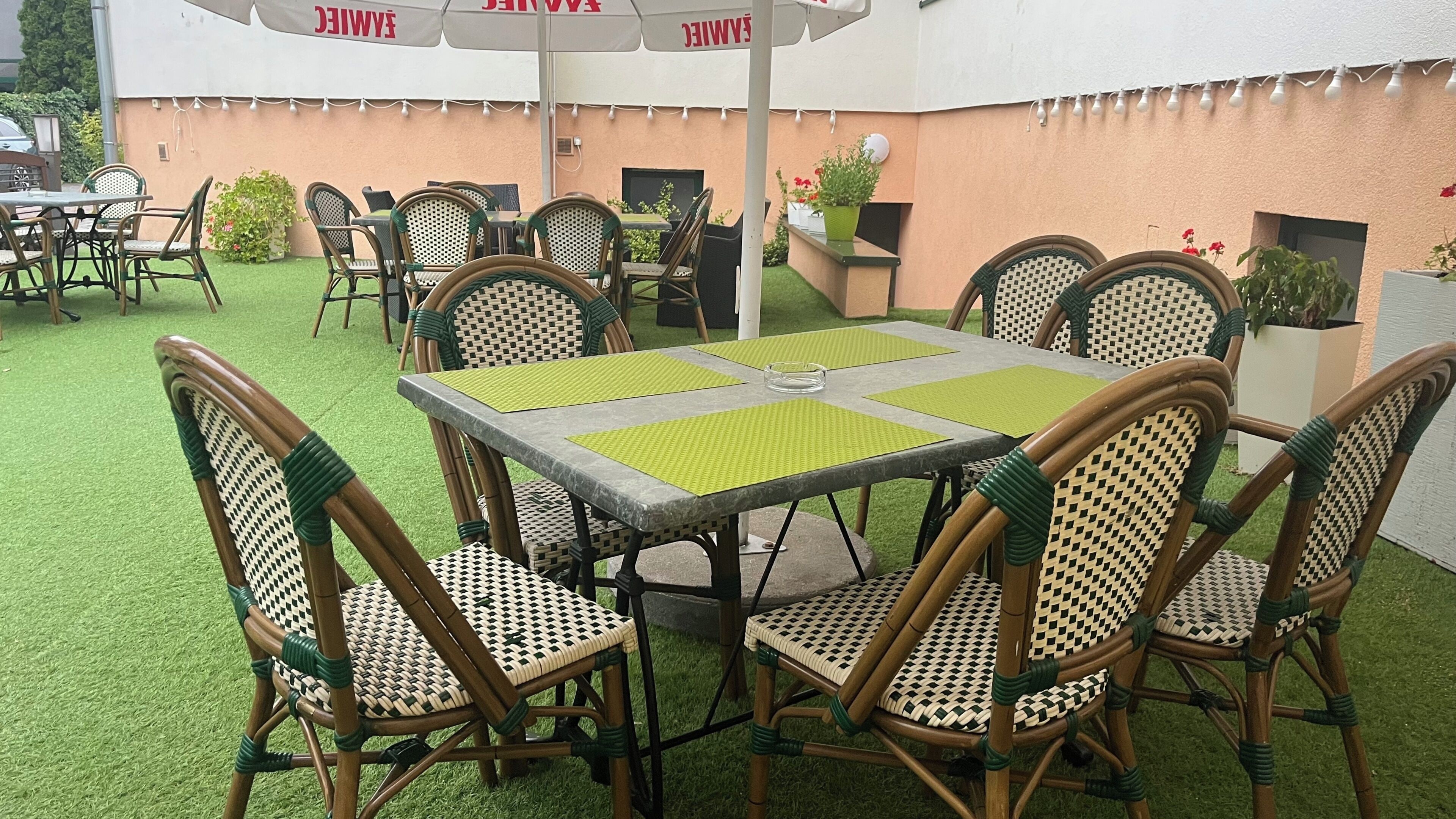 A patio area with outdoor seating, including tables and chairs, surrounded by greenery and a peach-colored wall.