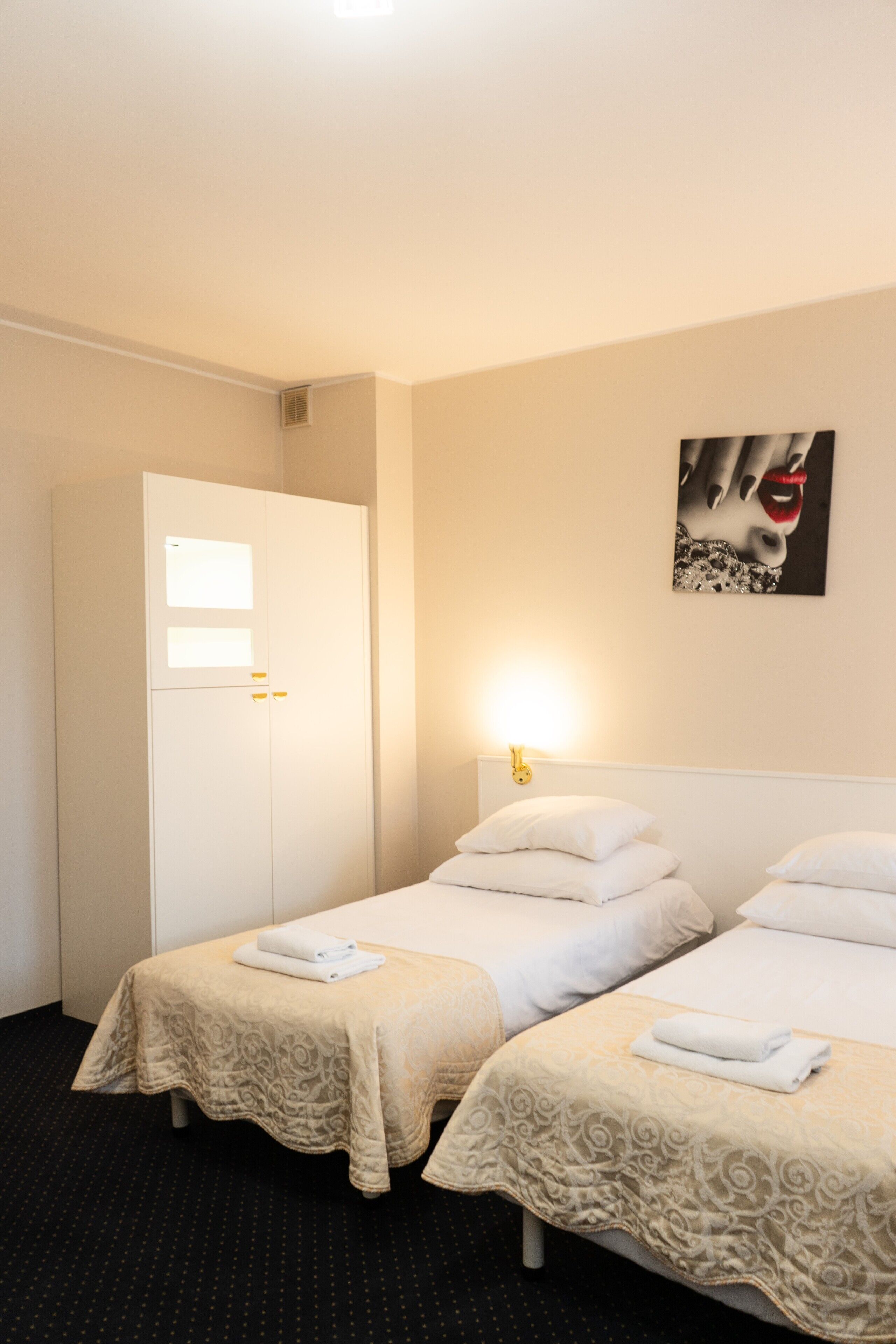 Ett hotellrum med två sängar, en garderob, en tavla på väggen och en lampa ovanför sängarna.