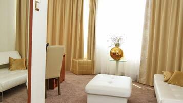 A modern hotel room with a white sofa, a matching ottoman, a glass coffee table with a golden vase, and beige curtains.