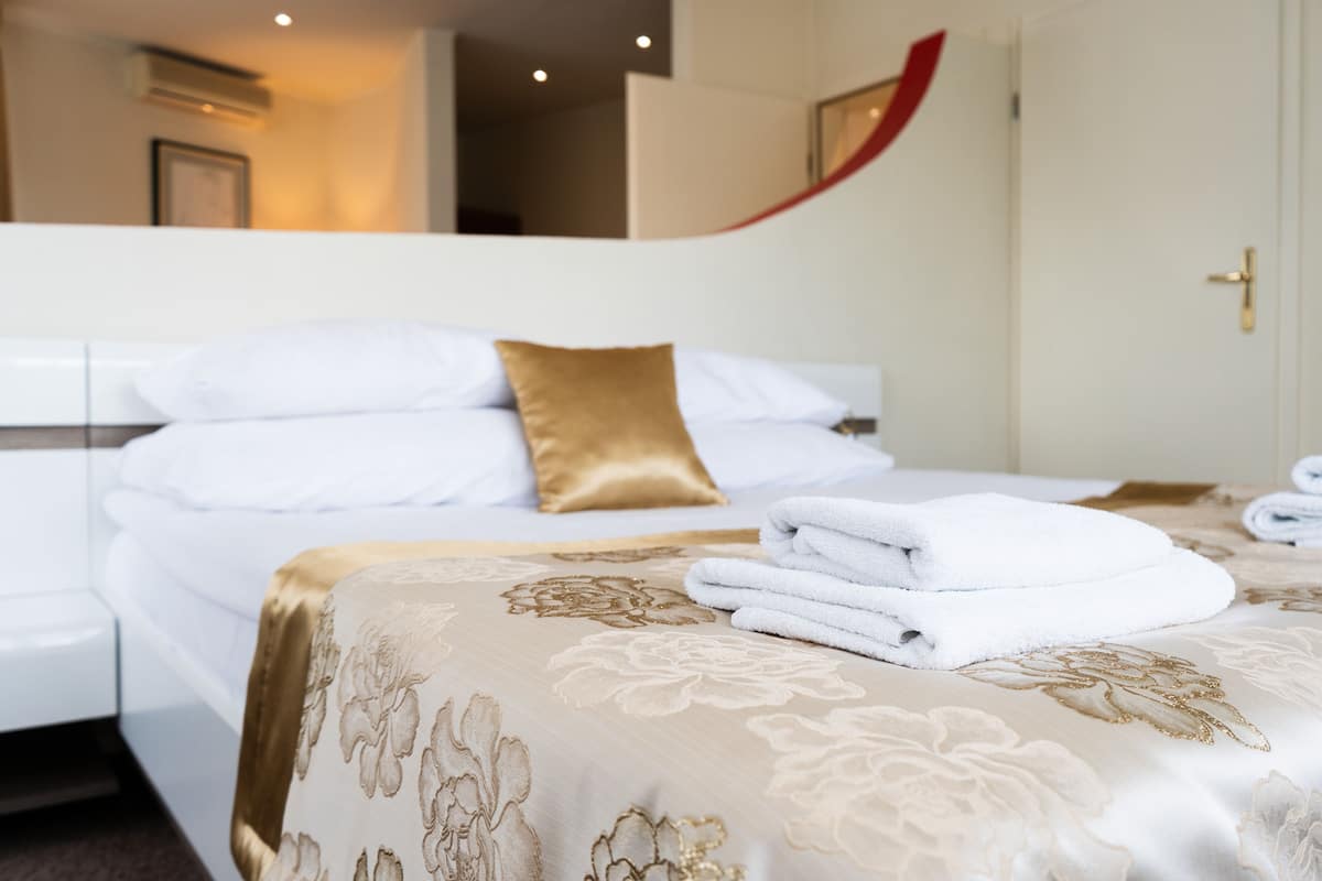 Una cama bien hecha con sábanas blancas y un almohadón dorado, una toalla blanca doblada y una colcha de motivos florales.
