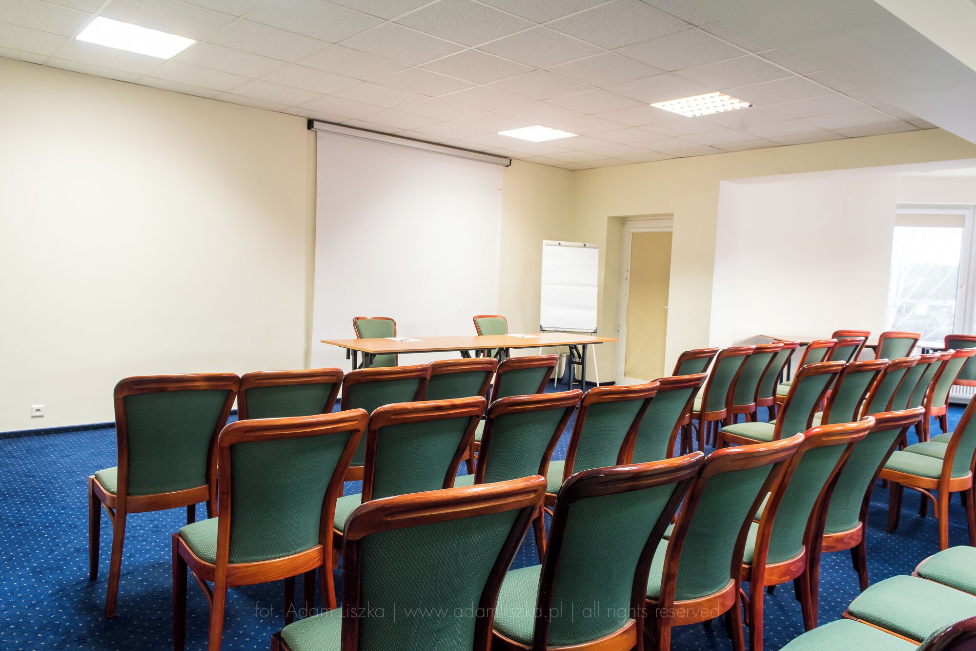 A conference room with a long table and rows of chairs, a whiteboard, and a projector screen.