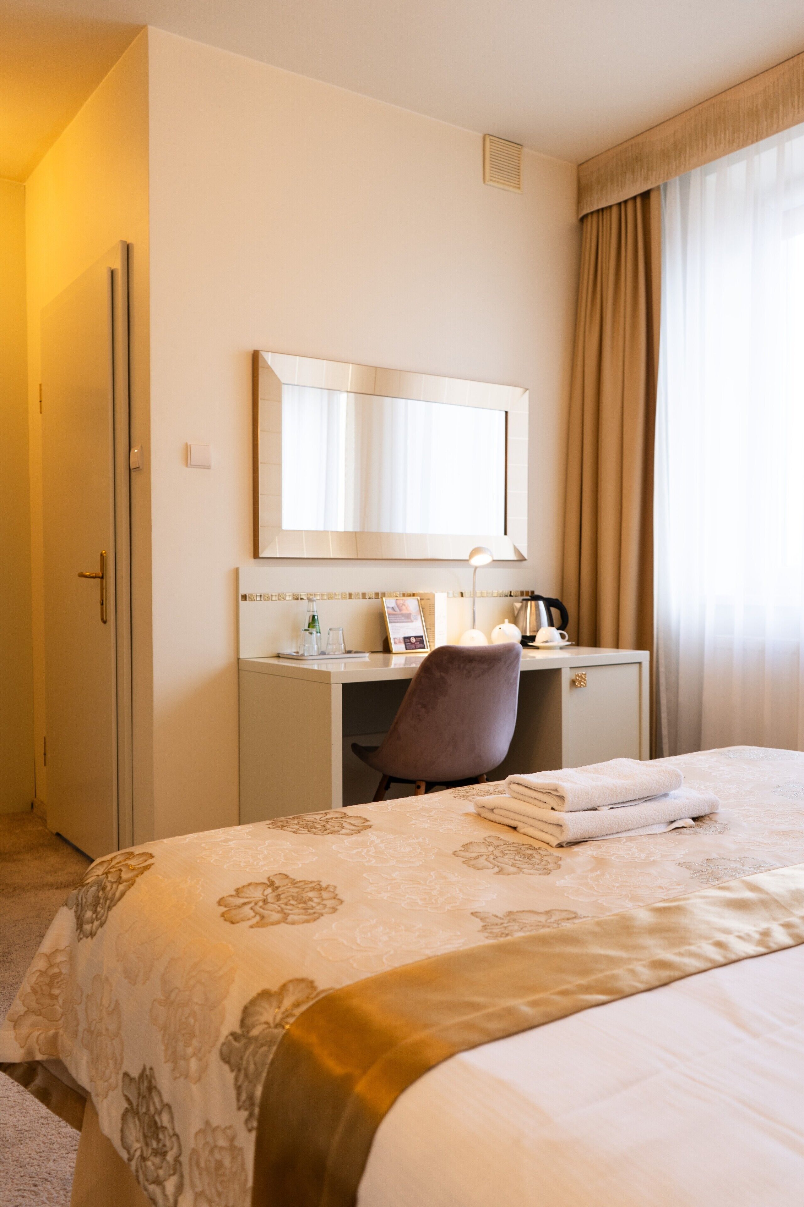 Ein Hotelzimmer mit einem Bett, einem Schreibtisch mit Stuhl, einem Spiegel und einem fensterbepflanzten Fenster.