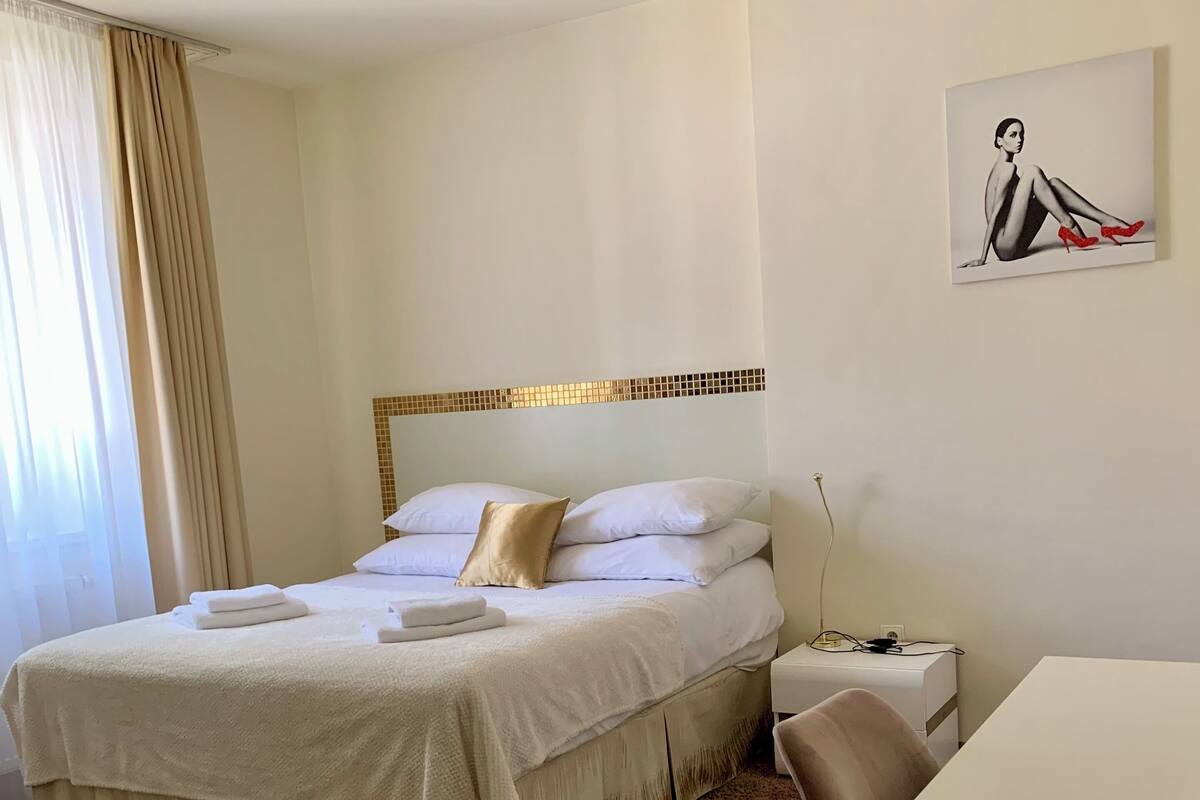 Habitación de hotel con cama, escritorio, silla, un cuadro en la pared y ventana con cortinas.