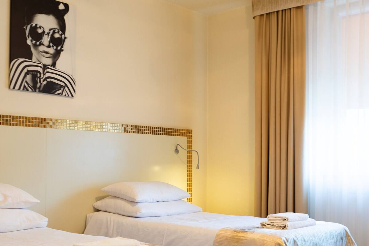 Habitación de hotel con dos camas, ropa de cama color crema, una lámpara fijada a la pared, una ventana con cortinas y un cartel en la pared.