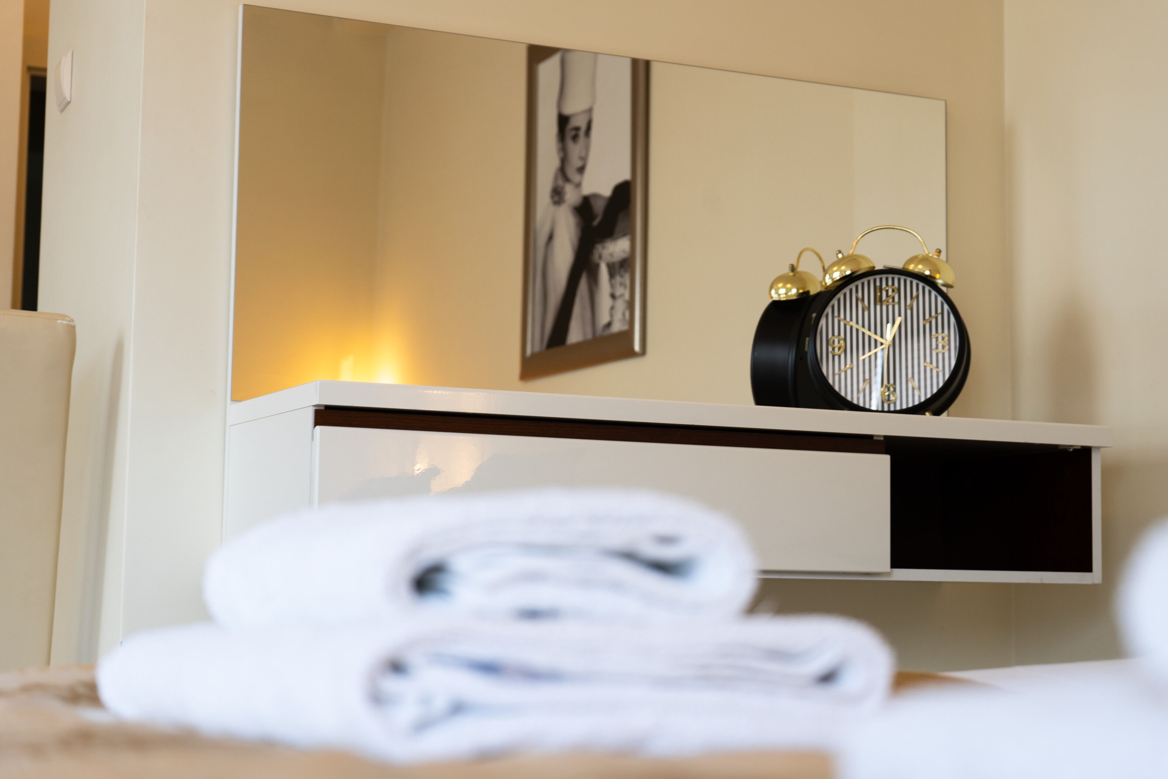 A neatly made bed with folded white towels, a black alarm clock, and a framed picture on the wall.