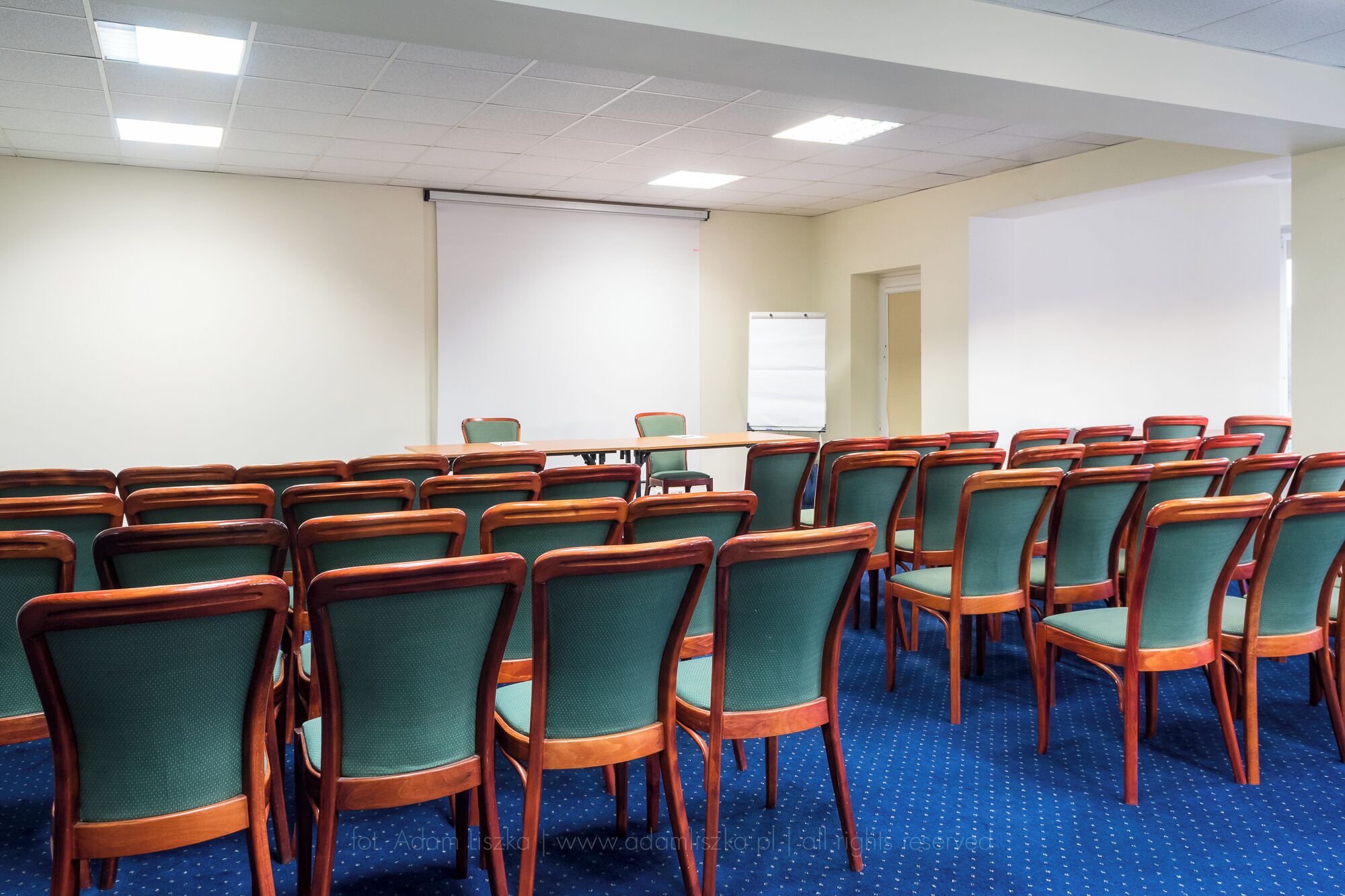 A conference room with rows of wooden chairs and a large table, whiteboard, and projector screen.