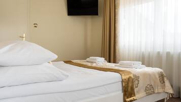 A neatly made bed with white linens and a gold throw blanket, a flat-screen TV mounted on the wall, and a window with sheer curtains.