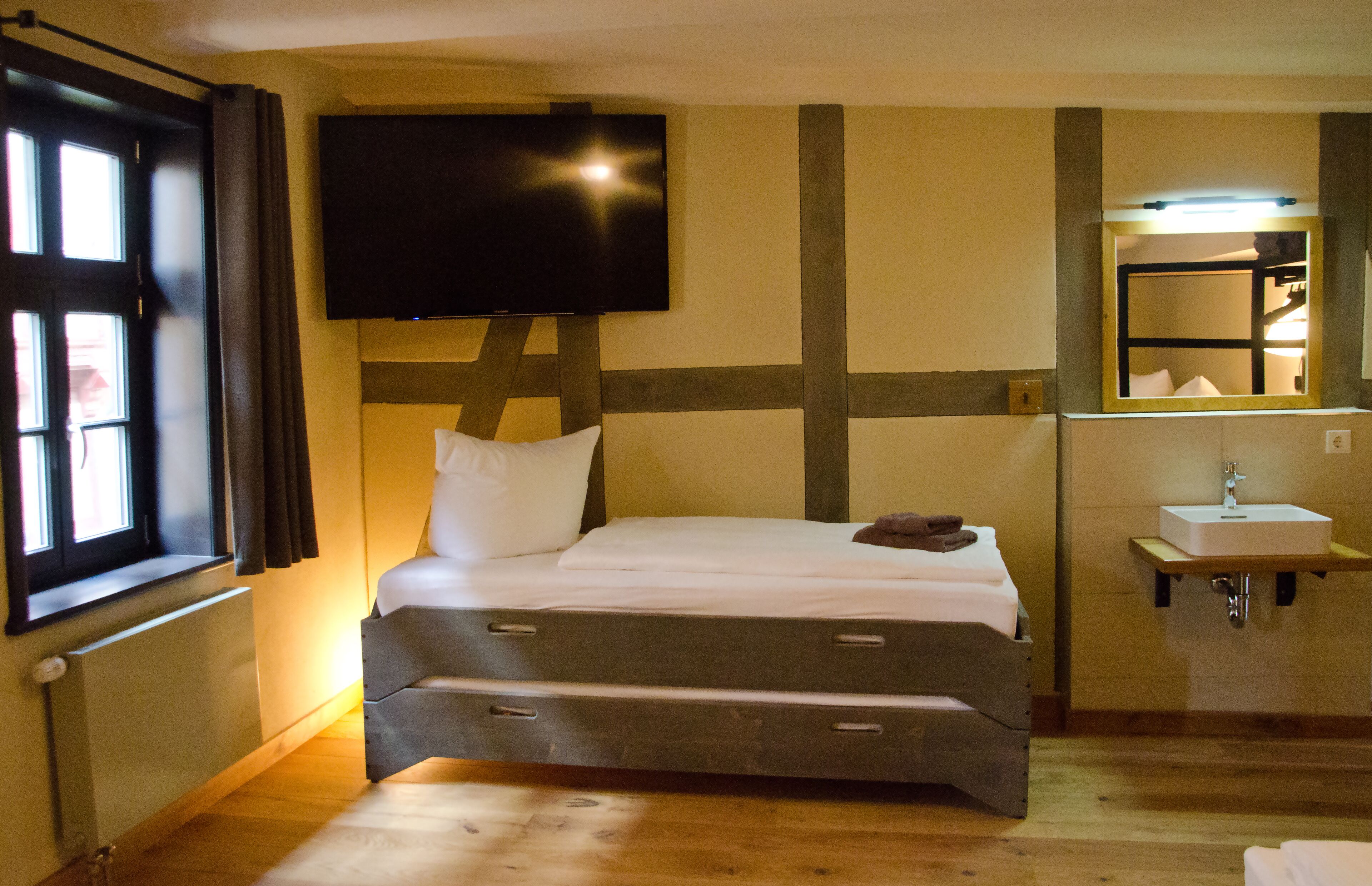 Un dormitorio con cama, televisor montado en la pared, ventana con cortinas y baño con lavabo y espejo.