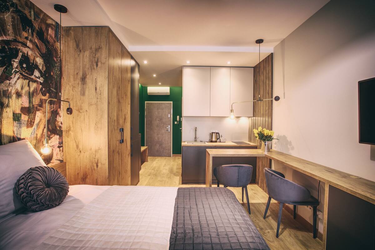 A bedroom with a bed, a desk, and a chair. There is a kitchenette with cabinets and a sink.