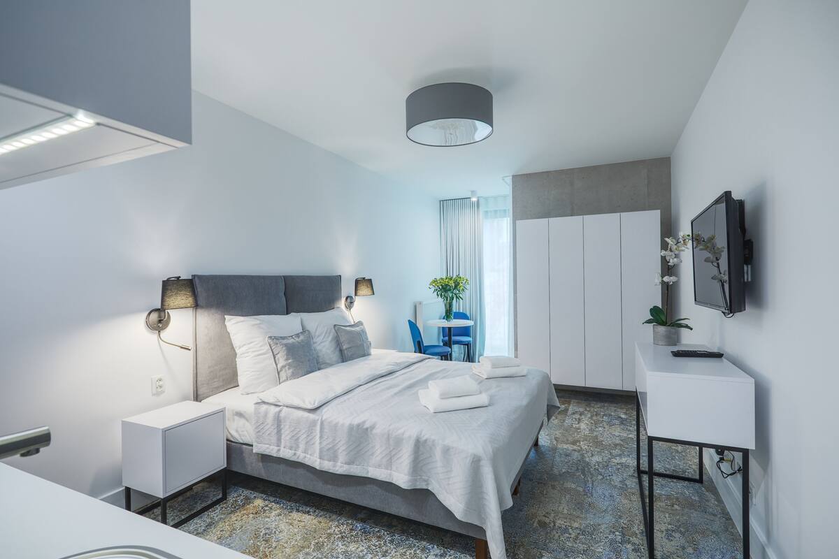 A modern hotel room with a large bed, bedside tables, a desk, a chair, a TV, and a window with curtains.