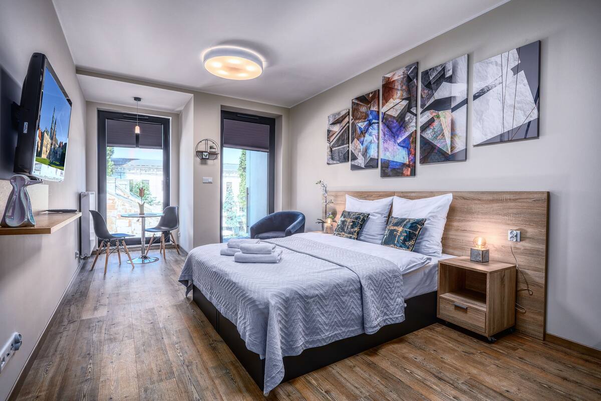 A modern bedroom with a large bed, a bedside table with a lamp, abstract wall art, and a view of the outdoors.