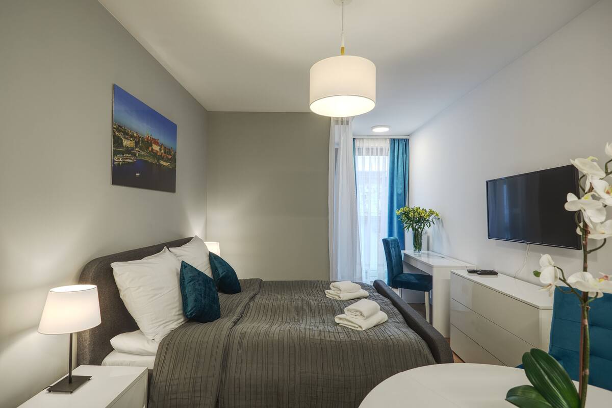 A hotel room with a bed, bedside tables, a desk, a TV, and a window with curtains.