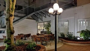 An indoor dining area with brick planters, wooden tables, and chairs. A staircase is visible in the background. - Adonis Lourdes Notre Dame De La Sarte (Lourdes)