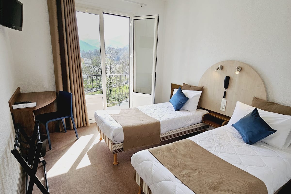 A hotel room with two beds, a desk, a chair, a TV, and a balcony with a view.