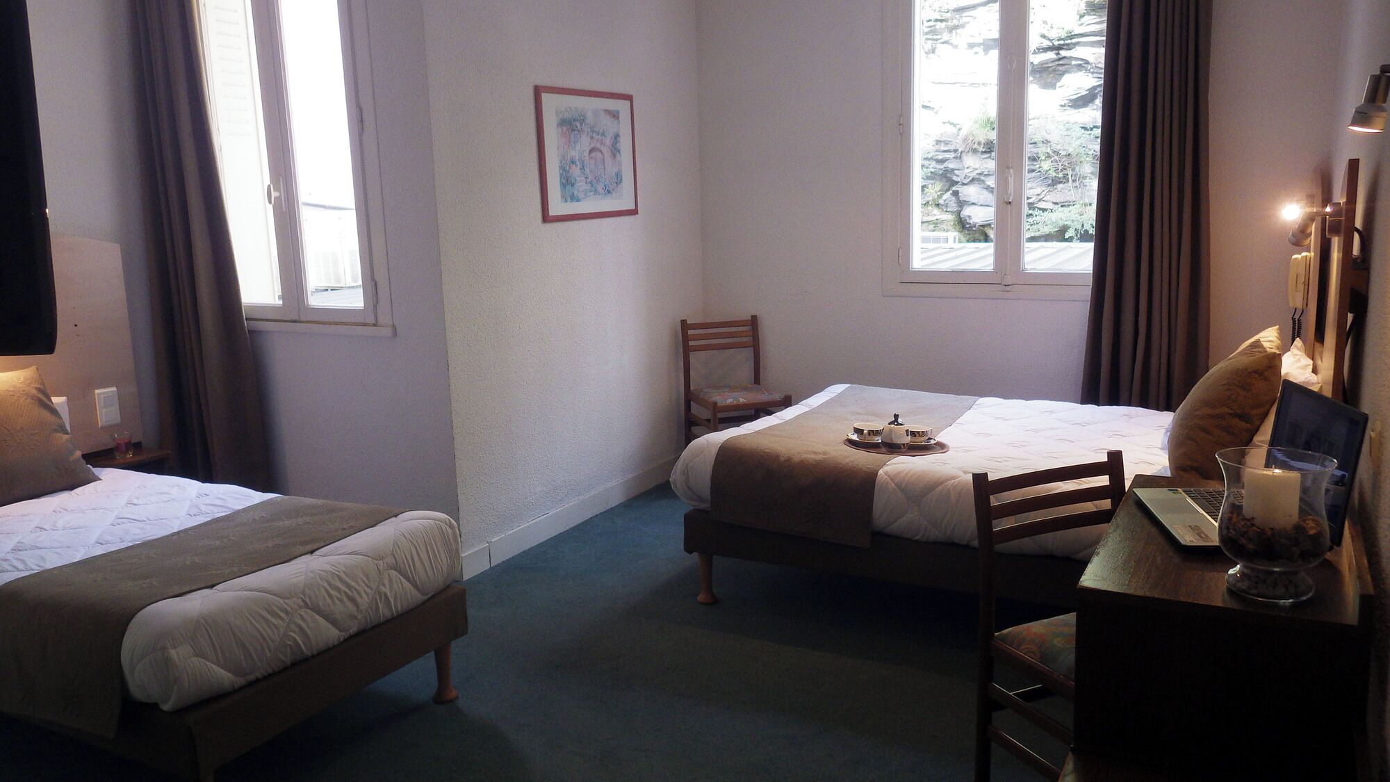 Camera d'albergo con due letti, una scrivania con un portatile, una sedia, un comodino con una lampada e una finestra con tende.