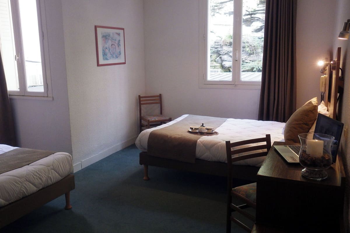 A hotel room with two beds, a desk with a laptop, a chair, a bedside table with a lamp, and a window with curtains.