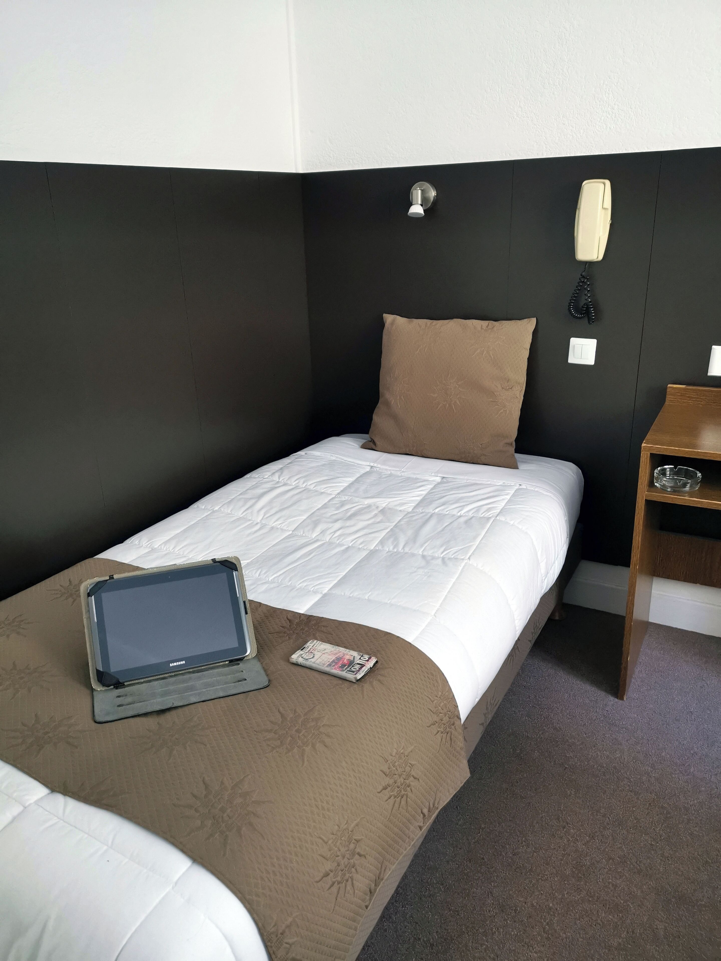 Camera d'albergo con un letto singolo, un comodino, un telefono e un tablet sul letto.